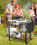Stainless Steel Pizza Oven Table For Ooni And Other Grills Heavy Duty Outdoor Grill Cart With Wheels And Handle 32 By 24 Inch Three Shelf Food Prep Cart For Griddle Movable Outdoor Pizza Cart shown in use at an outdoor gathering.