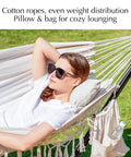 Woman relaxing in a handwoven tassel hammock with stand, pillows, and storage bag, set outdoors on grass. Beige oversized double hammock.