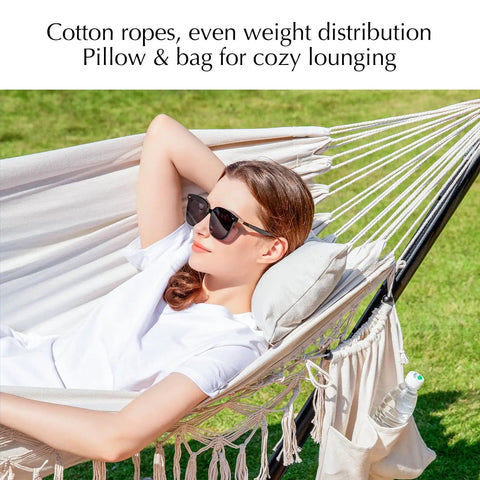 Woman relaxing in a handwoven tassel hammock with stand, pillows, and storage bag, set outdoors on grass. Beige oversized double hammock.