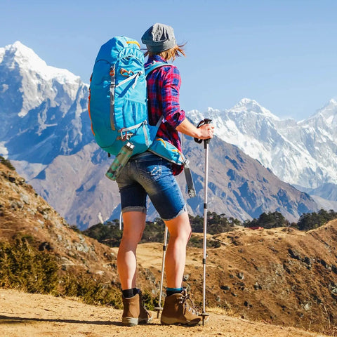 Hiker with a large blue backpack facing snow-capped mountains on a sunny day, wearing casual hiking attire, and holding trekking poles.