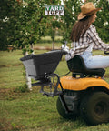 Person using Tow Behind 12V Lawn Tractor Spreader For Grass Seed And Fertilizer With 80 Pound Capacity And Lightweight Frame Weighing Sixteen And A Half Pounds For Easy Lawn Care And Even Distribution Across Large Yards attached to a tractor in an orchard.
