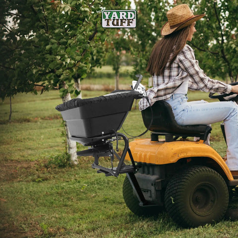 Person using Tow Behind 12V Lawn Tractor Spreader For Grass Seed And Fertilizer With 80 Pound Capacity And Lightweight Frame Weighing Sixteen And A Half Pounds For Easy Lawn Care And Even Distribution Across Large Yards attached to a tractor in an orchard.