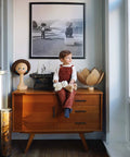 Child sitting on a wooden cabinet with decor items and a typewriter, framed black and white photo on wall.