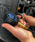 Close-up of JACO ElitePro digital tire pressure gauge with blue LCD, black ribbed housing and brass nozzle on tire valve.