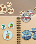 Premium 2.25 Button Maker Supplies Packed With Two Hundred Sleek Metal Badge Blanks And Crisp Paper Discs showing colorful custom pins on a notebook.