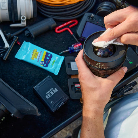 Close-up of hands wiping a camera lens with Invisible Glass lens cleaning wipes; blue 16-count package visible.
