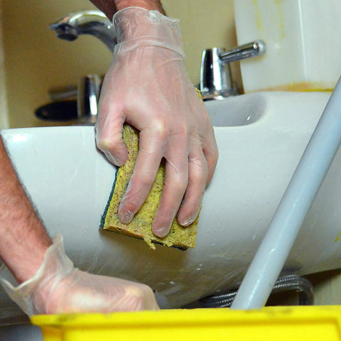 Person using Top Shelf Clear Vinyl Disposable Gloves straight up reliable 3 mil thickness for cleaning a sink with a sponge.