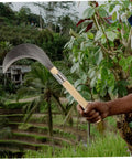 Long Handle Manganese Steel Grass Sickle For Weeding And Harvesting Garden Care With Non Slip Wooden Handle Comfortable Grip Versatile Workhorse Lawn Tool For Easy Yard Work And Maintenance in a lush garden setting.