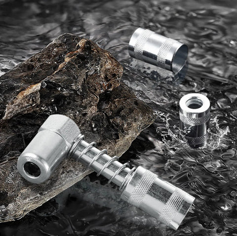 Angled front view of a silver 90-degree grease gun adapter with knurled grip and spring; fittings nearby on rock.