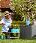 31 Gallon Weatherproof Resin Deck Box With Lid For Indoor Outdoor Storage Durable Grey Outdoor Storage Container next to woman gardening