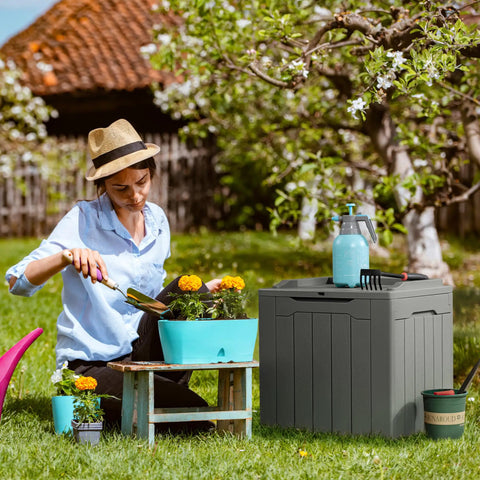 31 Gallon Weatherproof Resin Deck Box With Lid For Indoor Outdoor Storage Durable Grey Outdoor Storage Container next to woman gardening
