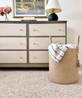 Tall Woven Laundry Basket for Living Room and Bedroom, Boho Farmhouse Round Wicker Blanket Basket for Towels and Clothes, Jute Laundry Hamper with Wooden Bead Tassel, Chic Home Decor 15 by 17.7 Inches next to a dresser with a lamp and flowers.