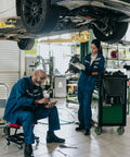 Mechanics working on a car in an auto repair shop, one seated with tools and the other standing with documents.