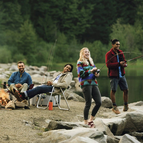 Group of friends fishing by a lakeside, sitting and standing with fishing rods, surrounded by rocks and trees, enjoying an outdoor activity.