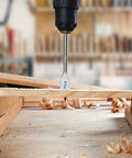 Front view of a steel spade drill bit drilling into wood, with wood shavings around.
