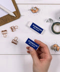 Hand holding a Mars Plastic Vinyl Erasers 4 Pack White Erasers For Clean And Precise Erasing Latex Free With Minimal Crumbling Great For Students And Artists Made In Peru, alongside office supplies on a desk.