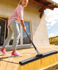 Real Deal Heavy Duty Push Broom With Long Handle Made To Crush Daily Grime On Smooth Floors Sweeps Sand Mud Leaves And Water Easy Without Breaking A Sweat In Bright Yellow 18 Inches Wide being used outdoors by a woman.