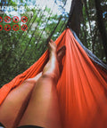 Person relaxing in an orange hammock with Camping Hammock Net With 360 Degree Mosquito Protection in a forest setting.