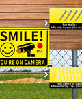 Smile You're On Camera Signs Two Pack Outdoor Video Surveillance Warning Signs 10x7 Rust Free Aluminum UV Printed Weatherproof Security Sign For Home Business Fence Driveway CCTV, mounted on a fence and wooden wall with installation options shown.