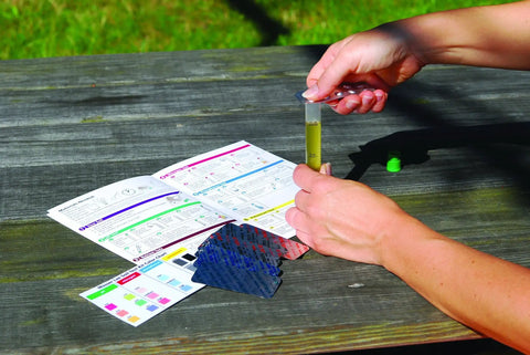 Person using ML1210 Soil Test Kit For Lawn, Garden And Potted Plants With 40 Tests, holding test tube with instructions on wooden table