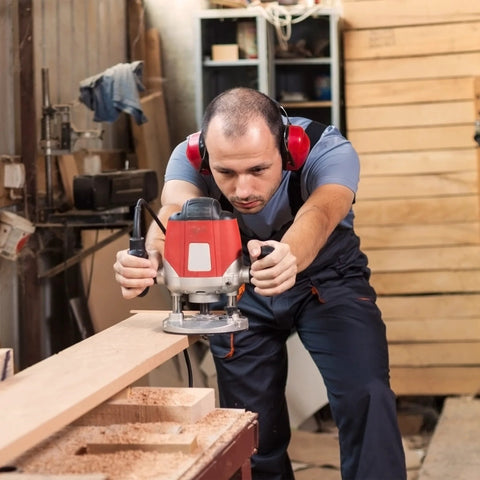 Red handheld router in use, guiding a wooden plank by a man wearing red ear protection in a woodworking shop.