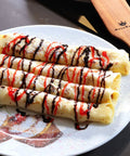 Four rolled crepes drizzled with chocolate and red syrup on a decorative plate, with a wooden kitchen turner visible in the background.