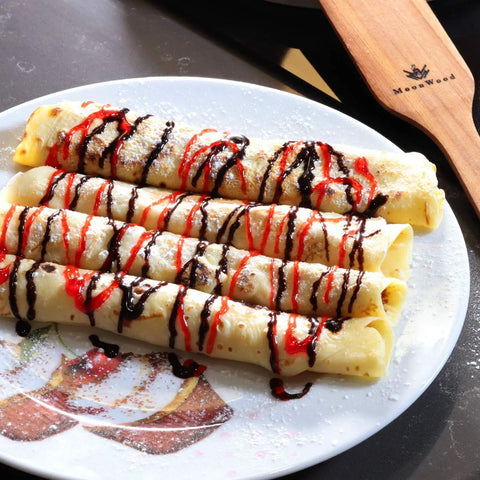 Four rolled crepes drizzled with chocolate and red syrup on a decorative plate, with a wooden kitchen turner visible in the background.