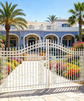 Ornate white gate in front of a villa with colorful garden and palm trees.