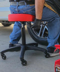 Rolling Shop Stool With Adjustable Height For Mechanics Padded Garage Seat With Smooth Rolling Wheels And ABEC Bearings Model 240250 Red in use next to a car wheel