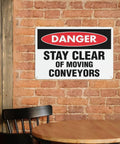 Stay Clear of Moving Conveyors Industrial Warning Sign 8 By 12 Inches Aluminum Wall Decor on brick wall near a wooden chair and table.