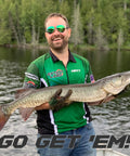 Man in green fishing jersey holding a large pike on a lake with trees in the background