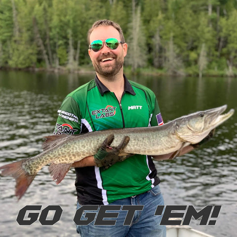 Man in green fishing jersey holding a large pike on a lake with trees in the background