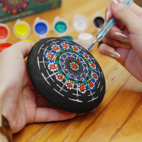 Person painting a mandala on a rock from Rock Painting Kit With Dotting Tools And Stencils And Acrylic Paints For Easy At Home Relaxing DIY Arts And Crafts Includes 5 Wooden Rocks 6 Mandala Stencils 12 Vibrant Colors And All The Dotting Tools For Adults And Women.