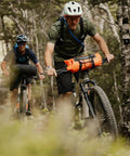 Cyclists using Spider Handlebar Cradle With 8L Heavy Duty Dry Bag For MTB Gravel And EMTB Bikes Quick Release Mount riding through forested trail.
