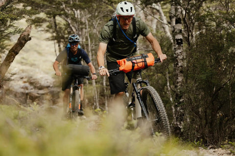 Cyclists using Spider Handlebar Cradle With 8L Heavy Duty Dry Bag For MTB Gravel And EMTB Bikes Quick Release Mount riding through forested trail.