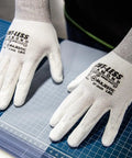 Close-up of hands wearing Cut-Less Dyneema Gloves With Polyurethane Palm, 1 Dozen LG, 13 Gauge Liner, laying on a cutting mat for precision.