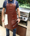 Person wearing Leather Grill Master Apron With Cross Back Straps Heat Resistant Full Grain Leather For BBQ Woodworking Barber And Welding Adjustable From M To 2XL Unisex Brown, standing by grill.