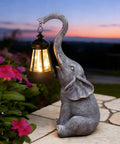 Elephant Statue Garden Decor With East Asian Charm featuring a solar lantern, warm gray finish, in a garden setting at sunset.