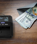 Portable Counterfeit Money Detector With LCD Display, Infrared And Magnetic Detection beside USD bills on a table.
