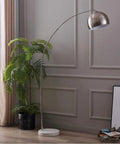 Arc Floor Lamp With Faux White Marble Base And Nickel Bell Shade For Living Rooms Offices Bedrooms Sleek Modern Mid Century Vibe Warm Ambient Task Lighting in a modern living room setting next to a potted plant.