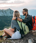 Two hikers with green and orange backpacks resting on a rocky mountain ridge.