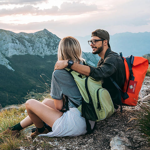 Two hikers with green and orange backpacks resting on a rocky mountain ridge.