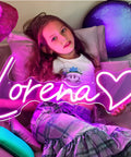 Pink 'Lorena' heart-shaped LED neon sign glowing beside a young girl in a bedroom with colorful decor.