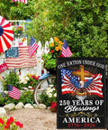 Patriotic 250th Anniversary USA Flag For Outside Garden Decor 12x18 Inch Double Sided We The People 1776 Celebration Flag For 4th Of July Outdoor Display in a garden setting with American flags and flowers.