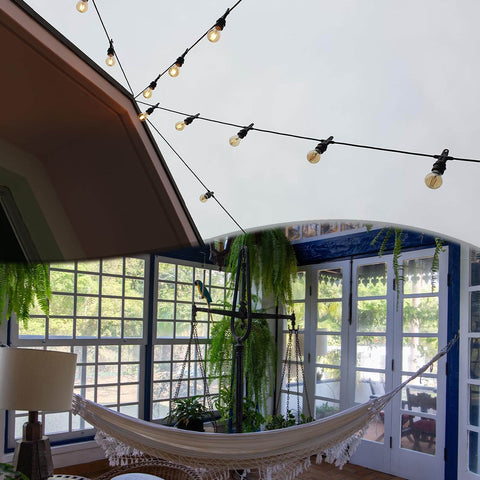Indoor sunroom with large windows, hammock, hanging plants, and string lights suspended from ceiling, creating a cozy atmosphere.
