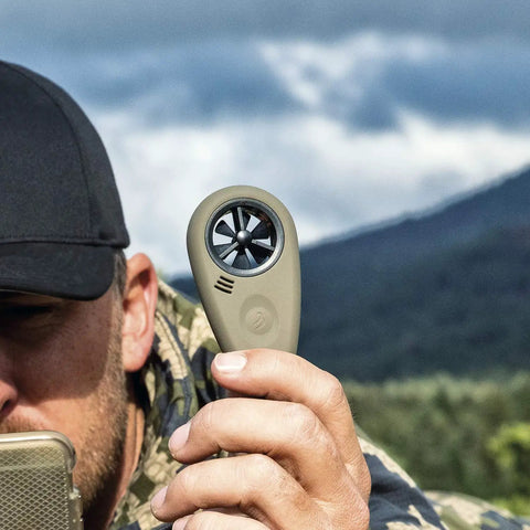 Person holding handheld wind meter with Bluetooth app connectivity for precision shooting and air flow measurement outdoors.
