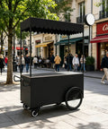 Kickass Heavy Duty Mobile Vendor Cart With Wide Shelter Canopy And Lockable Swivel Wheels Portable Black Food Display Stand in a busy street.