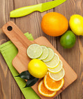 Wooden cutting board with sliced lemons, limes, and oranges next to whole citrus fruits and a green knife on a wooden surface.