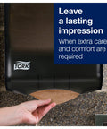 Tork multfold hand towel dispenser, black with natural towels; hand pulls a sheet, TORK logo visible.