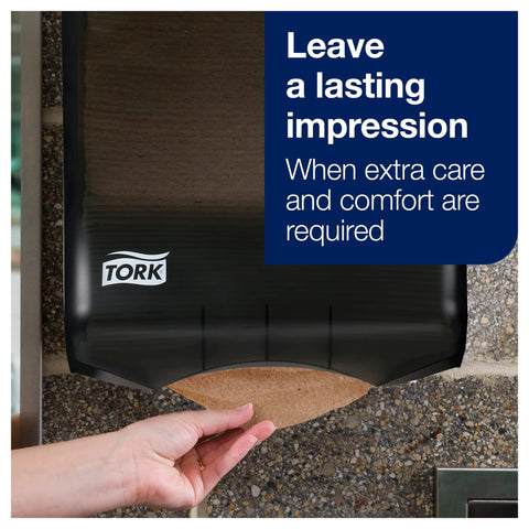Tork multfold hand towel dispenser, black with natural towels; hand pulls a sheet, TORK logo visible.
